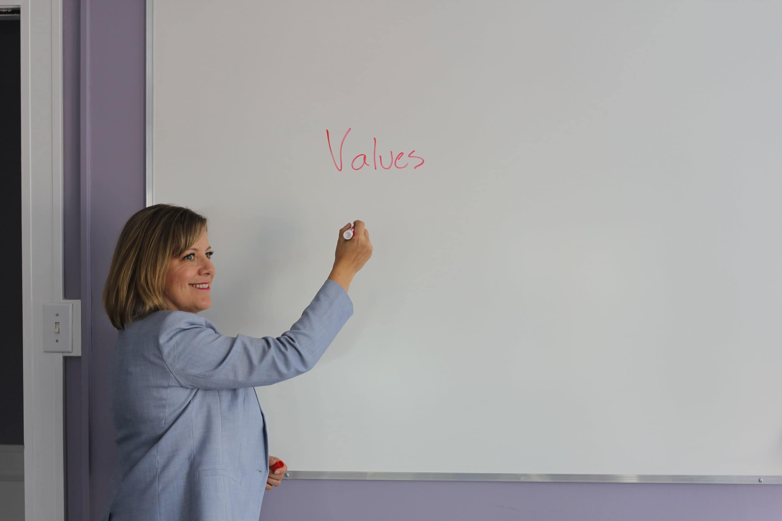 A midlife woman stands at whiteboard, marker in hand, helping women embrace a new chapter of purpose-driven leadership through her five years of Dare to Lead facilitation experience.
